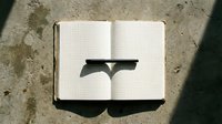 Open notebook with grid pages casting shadow on concrete surface, minimalist workspace setup for writing and planning projects.