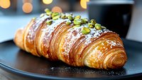 Fresh baked croissant with crushed pistachios and powdered sugar served on black plate against blurred bokeh background, close up view of flaky pastry.