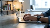 Professional businessman typing on laptop computer at modern office desk with warm lighting and contemporary workspace setup for remote work productivity.