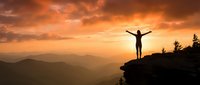 Person standing on mountain cliff with arms raised celebrating success at dramatic orange sunset with layered mountain ranges and evergreen trees.