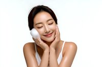 Young Asian woman with glowing skin applying facial cleanser foam with cotton pad, expressing joy during skincare routine on white background.