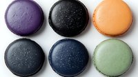 Six colorful macarons arranged in two rows featuring purple, black with sparkles, orange, gray, navy blue, and mint green flavors on white background.