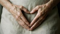 Elderly woman forming a heart shape with wrinkled hands on her stomach, symbolizing love, care, and health awareness.