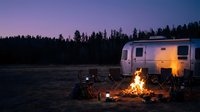 Campfire glowing beside a silver travel trailer at dusk, with forest silhouette and purple twilight sky in the background.