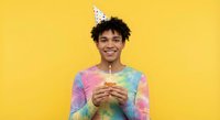 Happy young mixed-race man in party hat holding birthday cupcake with candle against bright yellow background for celebration designs.
