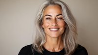 Confident mature woman with silver gray hair and natural makeup wearing black top smiling at camera against neutral background. Perfect for beauty and aging concepts.
