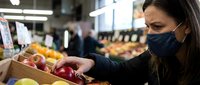 Woman wearing face mask shopping for fresh apples in grocery store produce section during pandemic safety measures for healthy lifestyle choices.