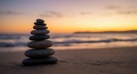 Balanced stone stack on beach at sunset with warm golden sky and gentle ocean waves creating peaceful meditation and wellness atmosphere.