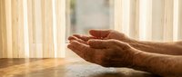 Elderly hands cupped in prayer or meditation gesture on wooden table with warm sunlight streaming through window blinds creating peaceful atmosphere.