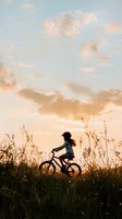 Silhouette of young girl riding bicycle through wildflower meadow at golden sunset, dramatic sky with soft clouds creates nostalgic summer mood.