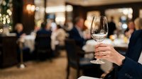 Empty wine glass held by person in formal attire at upscale dinner event with blurred guests and warm lighting in background, suitable for hospitality and event marketing.