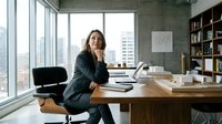 Young woman with dark hair and medium complexion, wearing a dark blazer, sitting at wooden desk in modern office with laptop, thinking pose, hand near face, high-rise window view, shelving, professional workspace for business, corporate, entrepreneur, productivity concepts.
