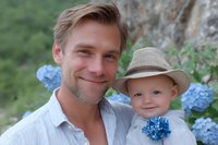Young Caucasian father holding smiling baby boy in straw hat against blue hydrangea garden background, tender family moment captured in soft natural lighting.