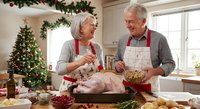 Senior Caucasian couple preparing Christmas turkey dinner together in festive kitchen with decorated tree, wearing matching red aprons and sharing joyful moment.