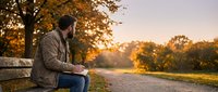 Young man sitting alone on wooden bench in autumn park during golden hour sunset, contemplating life with warm fall foliage and peaceful atmosphere.