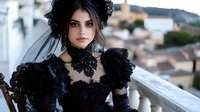 Young Hispanic bride in dramatic black lace wedding dress with curly hair and veil poses on vintage balcony against blurred cityscape background during sunset.
