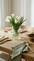 Fresh white tulips in glass vase on wooden dining table with linen napkins and wooden bowls creating elegant spring home decor atmosphere.