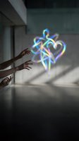 Hands reaching toward glowing holographic heart with rainbow light trails in modern interior space for digital love and connection concepts.