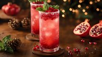 Red pomegranate cocktail with mint garnish on wooden coaster. Fresh fruit, ice cubes and sugar rim against dark background with festive bokeh lights.