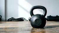 Black kettlebell weight on wooden floor in gym setting with resistance bands and training equipment, suitable for fitness, strength training, and workout content.