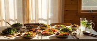 Rustic wooden table with an abundant spread of colorful healthy bowls, fresh salads, and grain dishes in warm natural sunlight.
