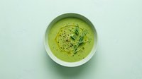 Fresh green cream soup in a white bowl, topped with microgreens, olive oil drizzle and black pepper on a light mint background.
