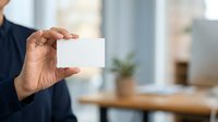 Man in dark business attire holding blank white card in modern office environment with wooden desk and potted plant, suitable for corporate branding and business communication materials.