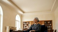 Mature woman with gray hair and light skin tone, wearing dark clothing and pearl necklace, sitting at desk in elegant home office with wooden bookshelves, arched window, and cream walls. Suitable for business, professional services, and lifestyle editorial use.