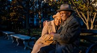 Happy senior African American couple embracing on park bench during golden hour sunset, showing love and togetherness in romantic outdoor setting.