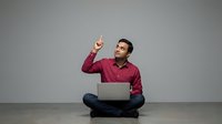 Young man with dark hair and olive complexion, wearing a burgundy button-up shirt, sitting cross-legged while using a laptop and pointing upward. Suitable for business, technology, education, and startup concepts.