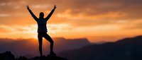 Silhouette of triumphant person with raised arms celebrating success on mountain peak at golden sunset with dramatic sky and mountain ranges in background.