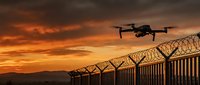 Drone flying over border fence at dramatic sunset with orange sky and mountains in background for security surveillance and monitoring operations.