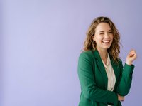 Young woman with wavy blonde hair, Caucasian ethnicity, wearing green blazer over white blouse, smiling confidently against purple background. Business professional portrait.