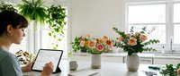 Young woman working on laptop in bright home office with fresh flowers and green plants creating productive workspace atmosphere.