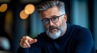 Mature businessman with gray beard and glasses pointing forward in modern office setting with warm bokeh lighting background.
