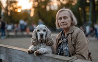 Senior Caucasian woman with blonde hair sitting in park at sunset with her cocker spaniel dog, both looking thoughtfully at camera.