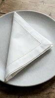 Folded white linen napkin with decorative stitching placed on neutral ceramic plate, styled on rustic wooden surface for dining and table setting concepts.