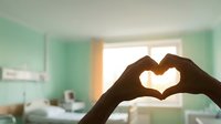 Hands forming heart shape silhouette against bright window in modern mint green bedroom interior for love, romance and home comfort concepts.