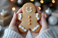 Hands holding festive gingerbread cookie with white icing smile and decorative buttons against warm Christmas lights background for holiday baking concepts.