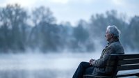 Senior Caucasian man with gray hair sitting alone on wooden bench by misty lake during peaceful morning contemplating nature in serene outdoor setting.