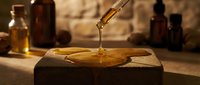 Liquid amber or essential oil dripping onto wooden surface with glass bottles in warm, blurred background for aromatherapy and wellness product use.