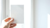 Caucasian woman's hand holding blank white card or paper against bright window light, suitable for mockups, presentations, and marketing materials.