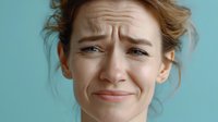 Young woman with concerned facial expression showing worry, anxiety, or discomfort against turquoise background, conveying emotional distress.