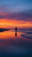 Silhouette of fisherman casting line on beach during dramatic sunset with vibrant orange and purple sky reflecting on wet sand and ocean waves.