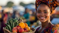 Young African woman vendor smiling at outdoor market with fresh tropical fruits in wicker basket wearing colorful traditional headwrap and clothing.