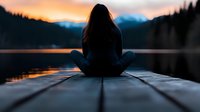Young woman in lotus position on wooden pier at sunset, silhouetted against mountain lake with orange sky and forest reflections, practicing meditation and mindfulness.