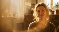 Middle-aged Caucasian woman smiling warmly in cozy home interior with golden sunlight streaming through windows creating peaceful domestic atmosphere.