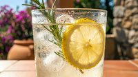 Refreshing iced lemonade with rosemary sprig and lemon slice in glass, sparkling summer drink outdoors on sunny patio.