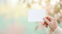 Hand holding blank white card against soft blurred spring garden background with blooming flowers, ideal for greeting cards, invitations, and promotional messaging.