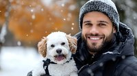 Young bearded man in winter jacket and knit hat smiling with small white poodle dog outdoors in snowy weather during cold season.